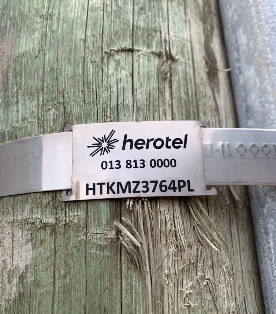 The photo is close up of a weathered wooden pole wrapped with a galvanized metal strapping. Affixed to that small white plastic tag. The tag carries a black sunburst style logo and the word herotel along with a telephone and pole number.
